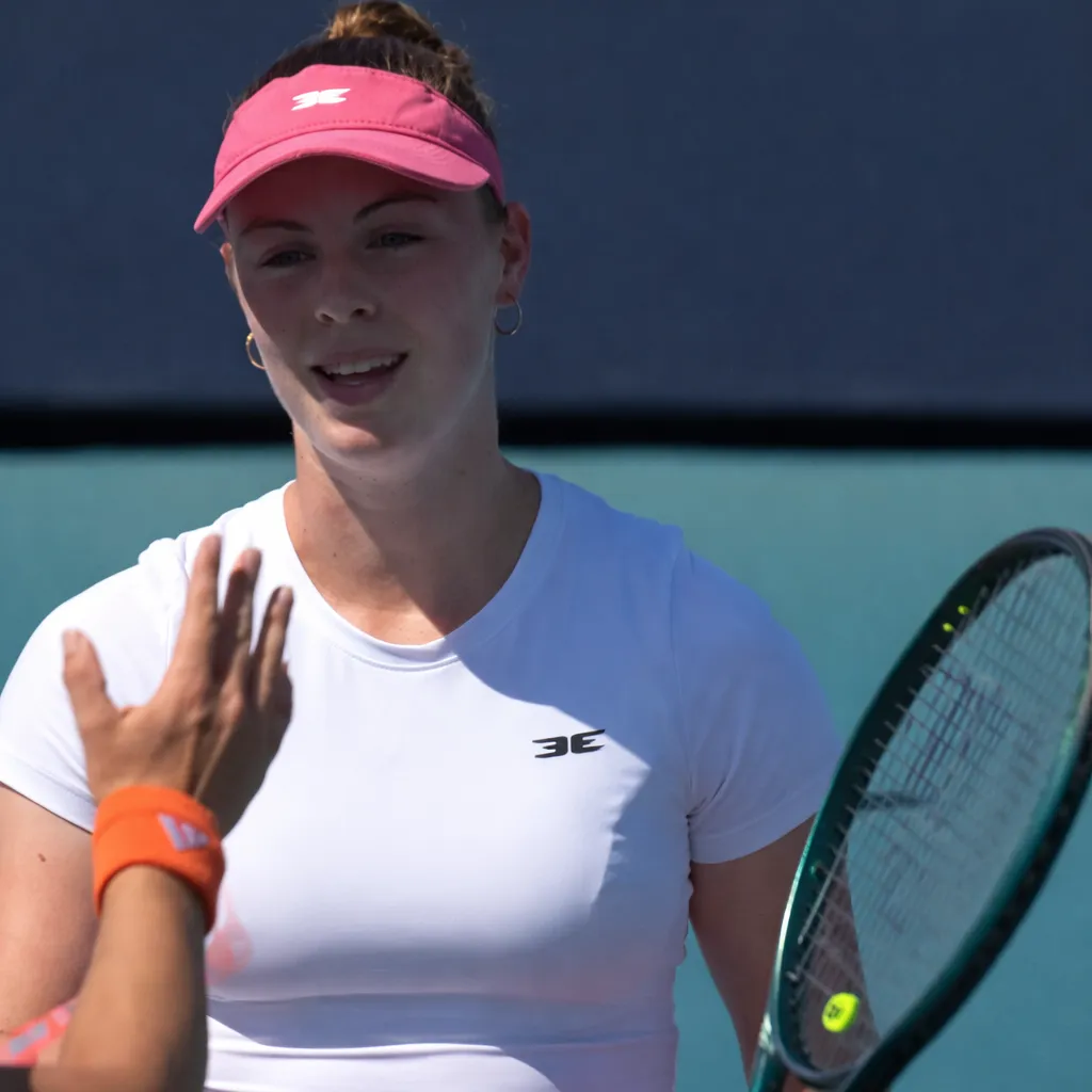 Talia Gibson speaks at the net during a match, holding her racket while interacting with her opponent on court.