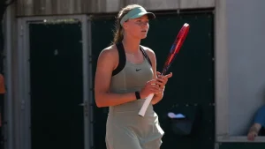 Teenage tennis player Alina Korneeva standing on a red clay court holding her racket, wearing a light green Nike outfit and visor